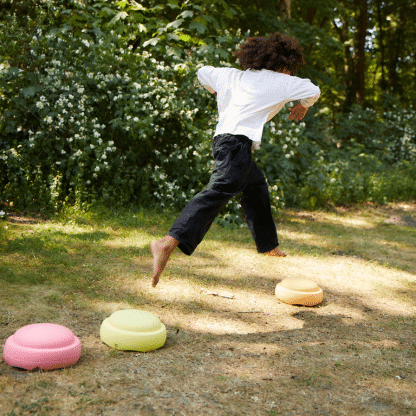 kind met zwarte broek en witte tshirt die aan het springen is over 3 stapelstein stenen in pastel roze, geel en oranje. de stenen liggen op een aarde weg langs een groen bos